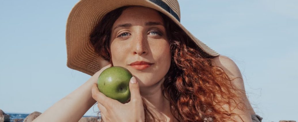 A relaxed woman with a hat holds a green apple, enjoying a sunny beach day.