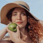A relaxed woman with a hat holds a green apple, enjoying a sunny beach day.