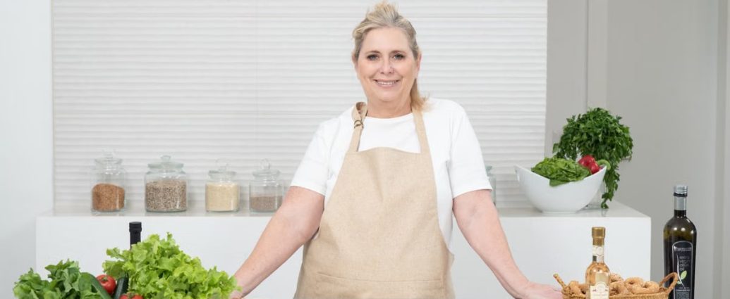 A mature woman in an apron stands at a kitchen counter with fresh produce and cheese, ready for cooking.