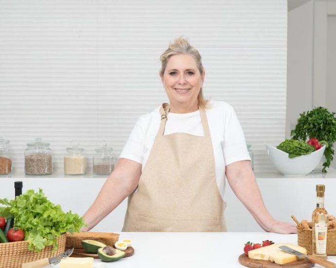 A mature woman in an apron stands at a kitchen counter with fresh produce and cheese, ready for cooking.